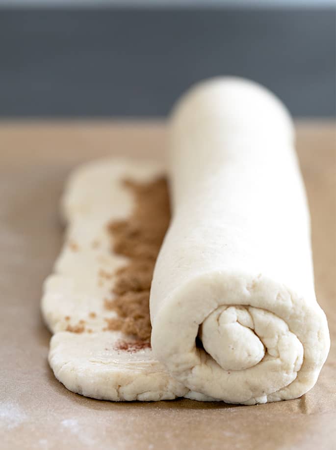 a partially coiled white rectangular piece of gluten free cinnamon roll dough with brown sugar and dark brown cinnamon showing on the uncoiled part, all on brown paper