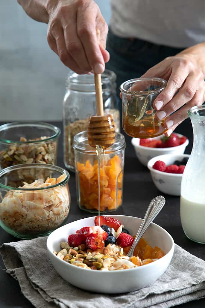 A person holding a jar of gluten free honey and drizzling it into the bowl with red and blue berries and muesli and a metal spoon on a tan cloth