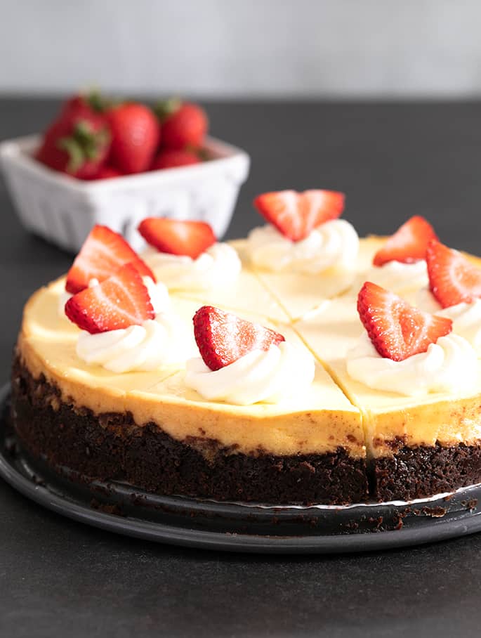 whole gluten free cheesecake with dark brown cookie crust sliced into 8 triangles each with a dollop of white whipped cream and a strawberry slice