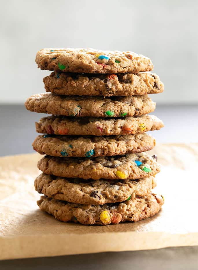 Stack of 8 light brown round gluten free monster cookies with colored candy pieces on brown paper