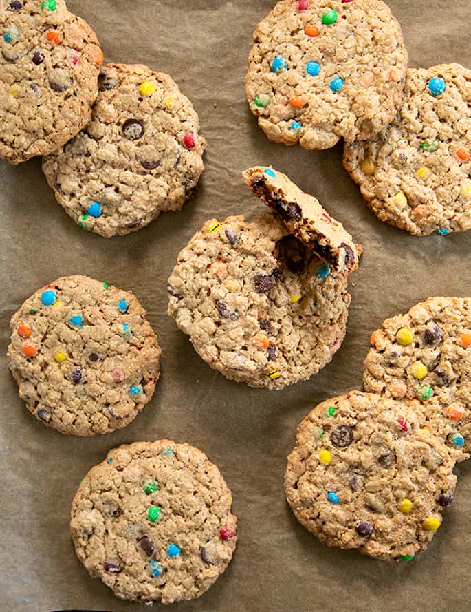 Overhead image of light brown round cookies with colored candy pieces baked in on brown paper and one cookie broken. 