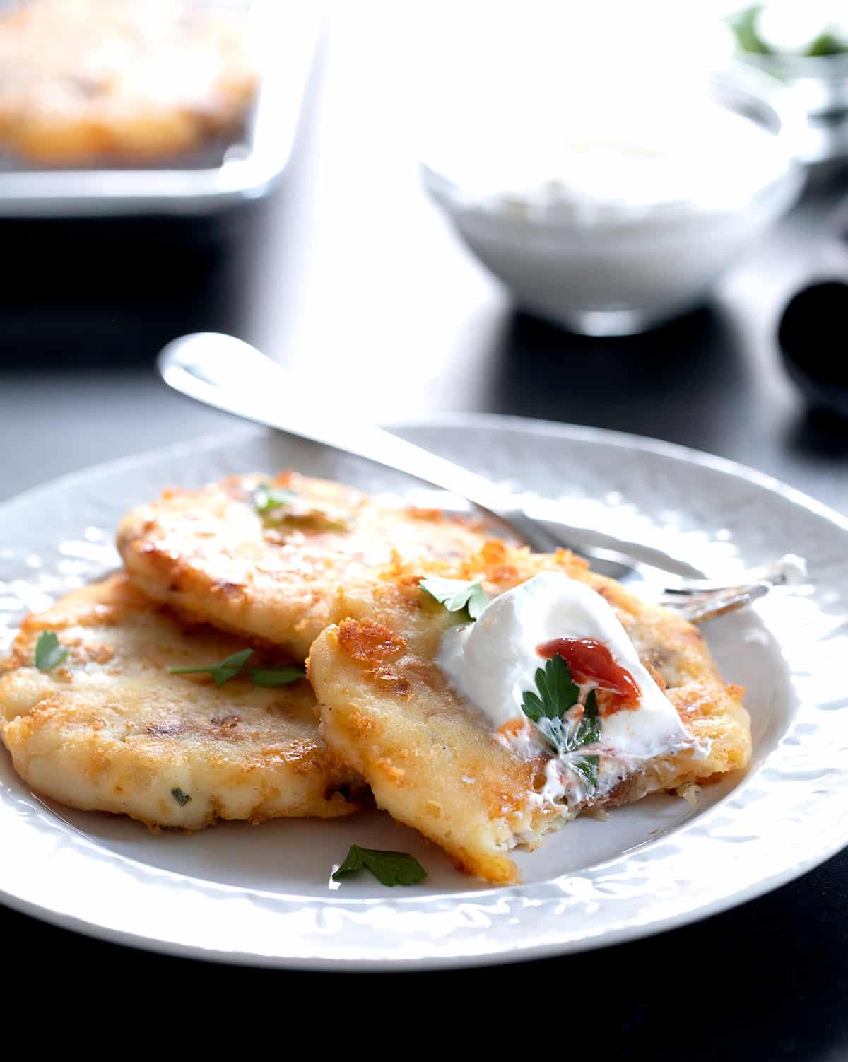 Three cakes of fried mashed potatoes on small white plate with sour cream and parsley leaves