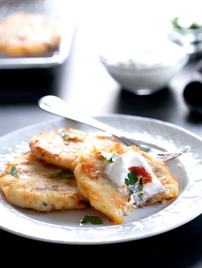 Three cakes of fried mashed potatoes on small white plate with sour cream and parsley leaves