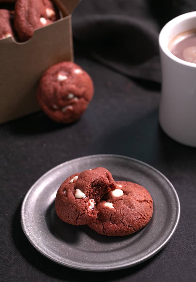 Two red round drop cookies with white chocolate chips on a small round metal plate with a bite missing from one cookie