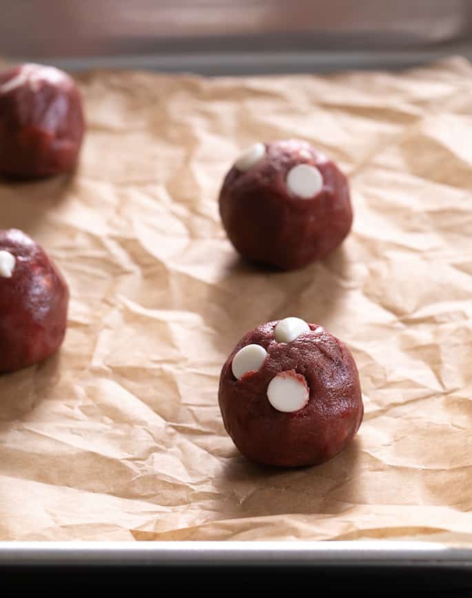 Raw round balls of red cookie dough with white chocolate chips on top on wrinkled brown paper on metal rimmed baking sheet