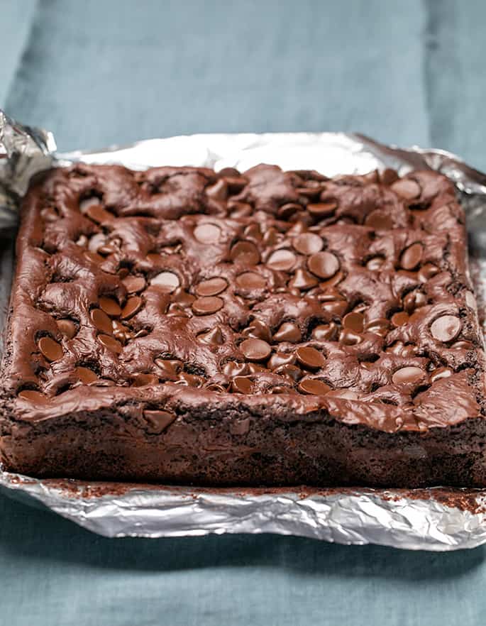 Baked uncut brown bars with chocolate chips baked on top, resting on foil that's been peeled away, all on blue cloth