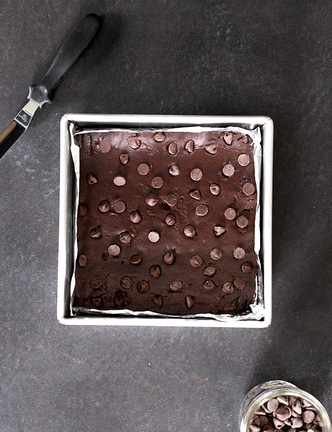 Overhead image of raw brownie batter in lined square metal baking dish with small offset spatula next to the pan