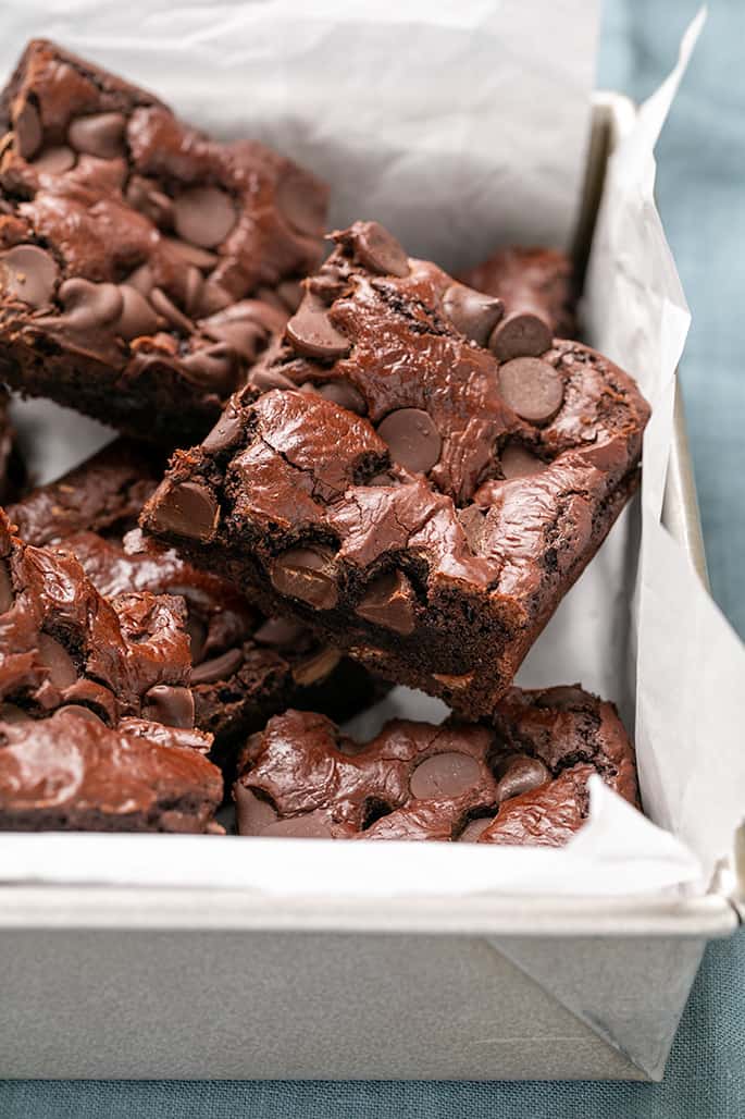 Square brown bars with chocolate chips on top piled in a square metal baking pan lined with white paper, all on blue cloth