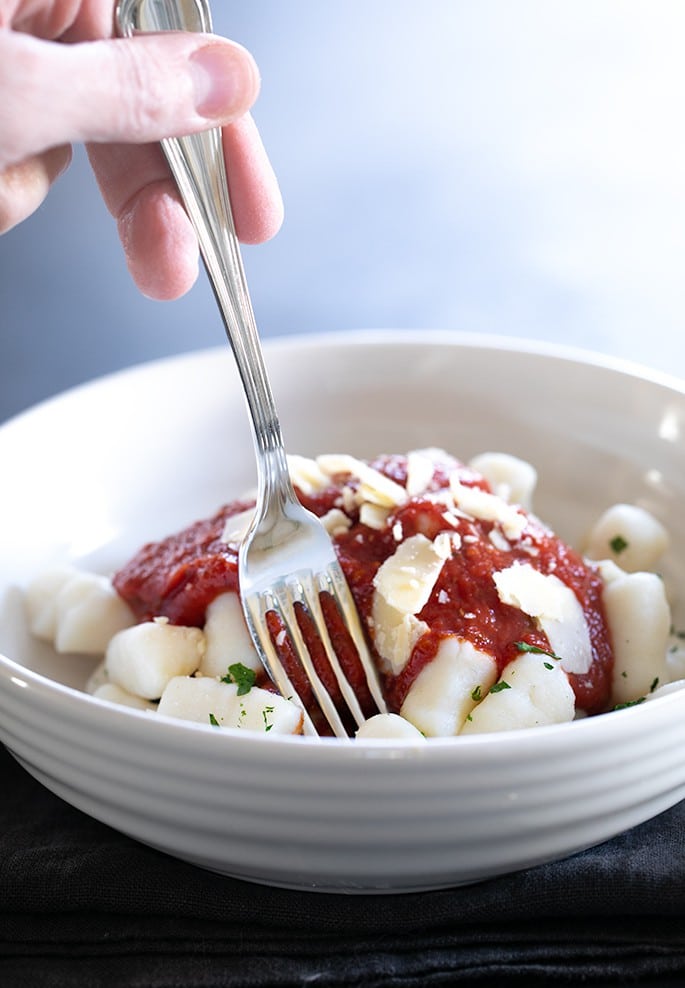 fingers holding a fork in a round bowl of dumplings with tomato sauce and cheese shavings