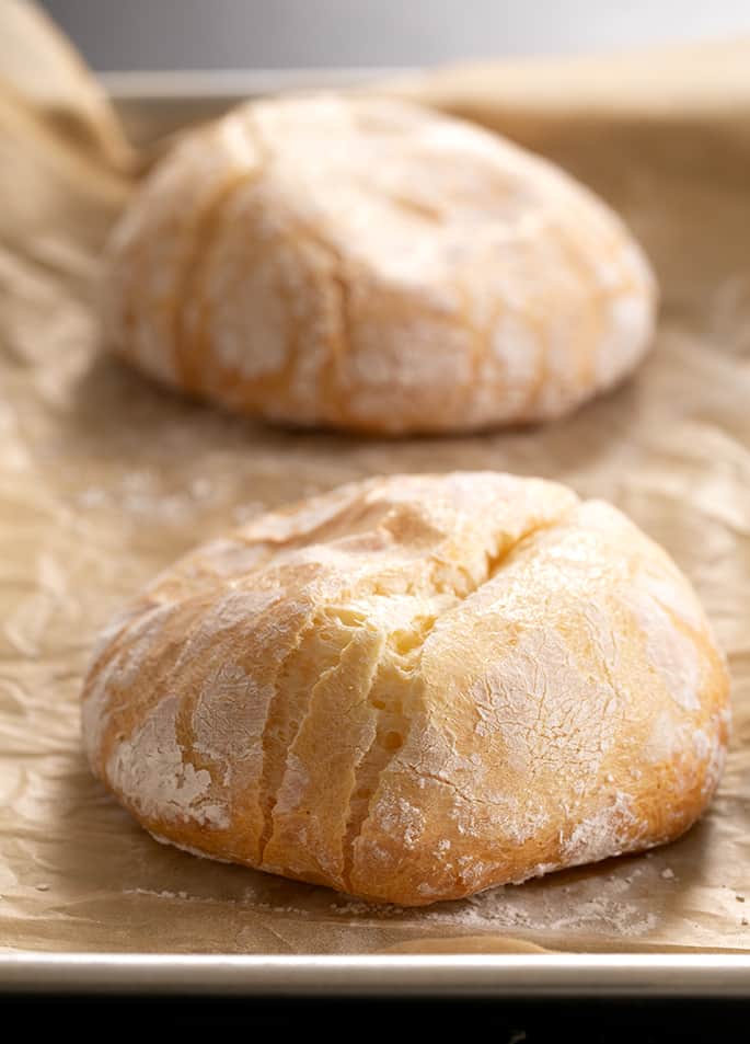 Light brown rounds of baked bread with cracks in the crust on wrinkled brown paper on a metal baking tray