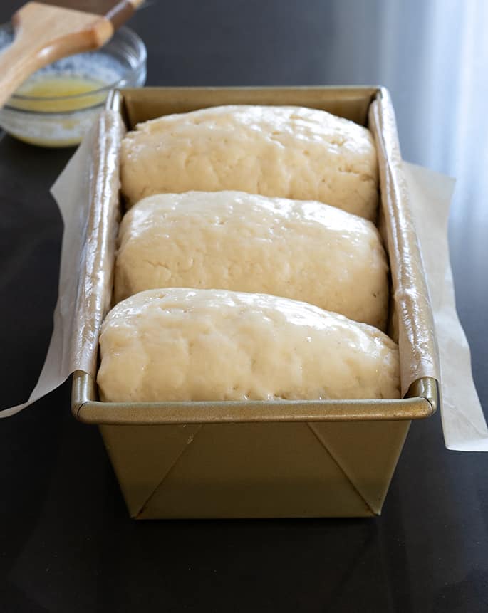 A loaf pan with white paper lining and three pieces of raw bread dough