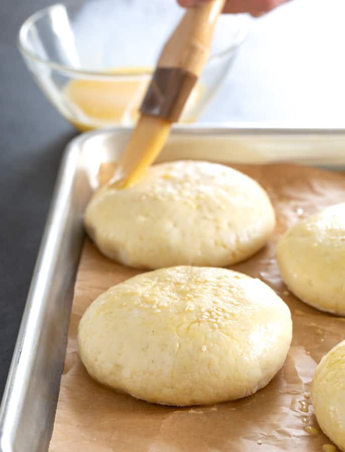 Pastry brush painting egg wash on raw round buns with sesame seeds on brown paper on baking tray