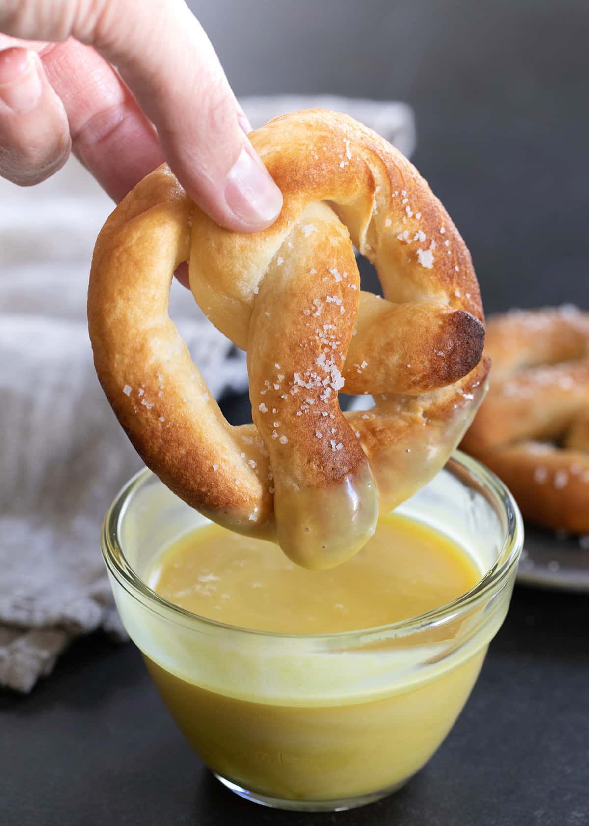 Fingers holding a light brown soft pretzel with white flaky salt and coated in yellow honey mustard on bottom