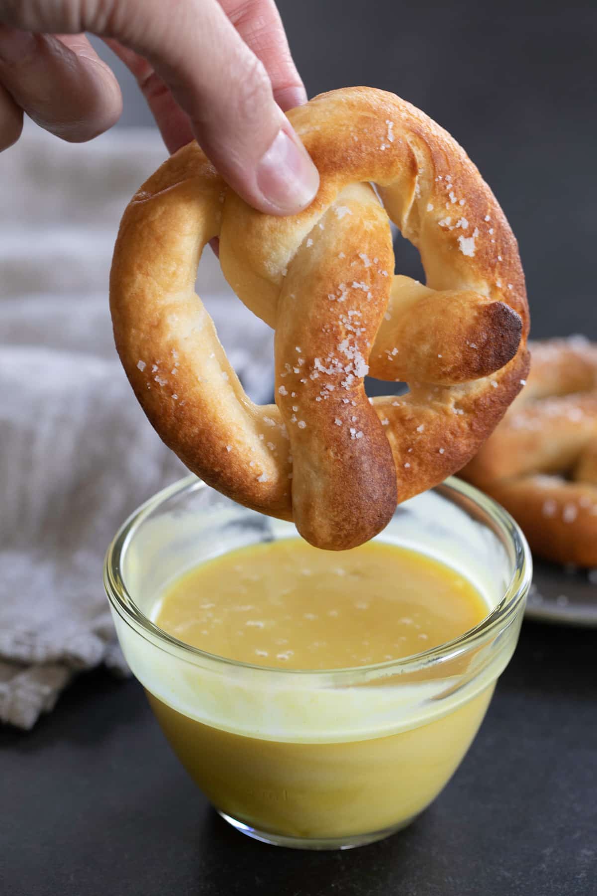 fingers holding a light brown gluten free soft pretzel with flaky salt above a small bowl of light yellow honey mustard