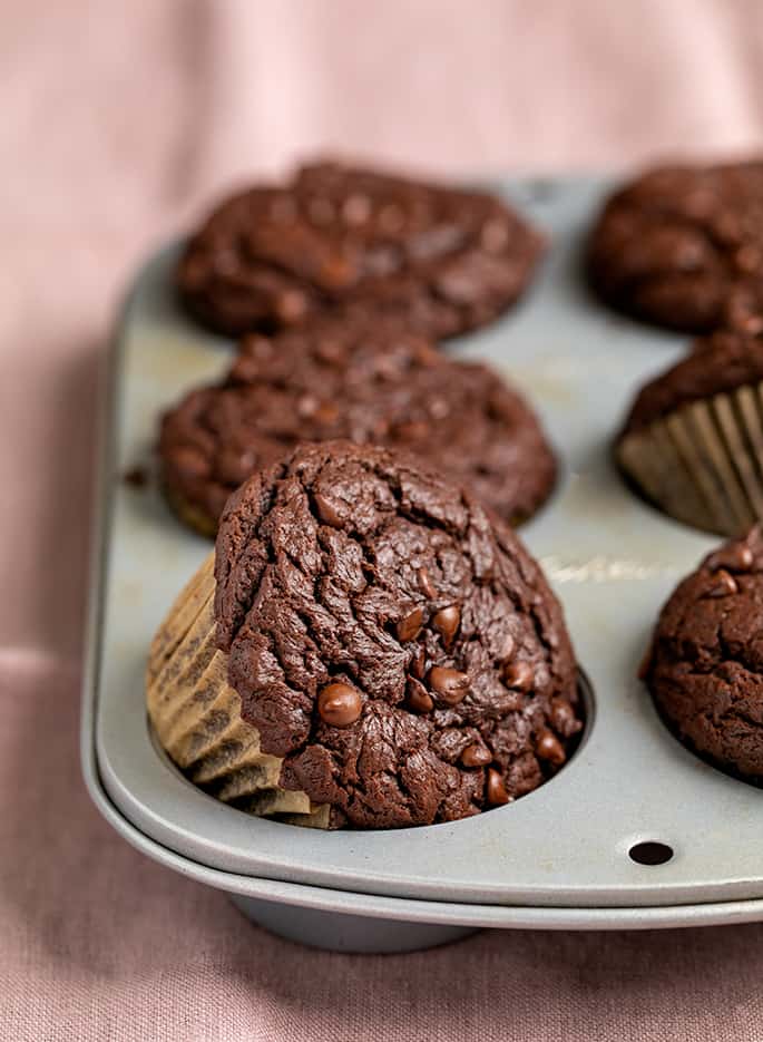 Brown chocolate chip muffins in liners in 6-cup metal muffin tin on pink cloth