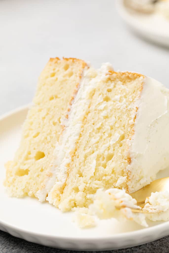 Closeup image of one slice of vanilla layer cake with white frosting with fork with crumbs on small white plate