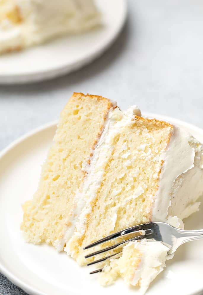 One slice of vanilla layer cake with white frosting in center on white plate with fork in corner of slice