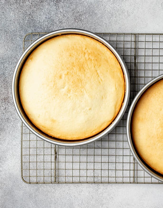 Overhead image of 2 yellow cakes in round metal baking pans on wire rack