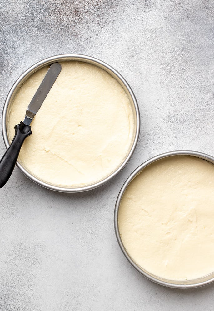 Two round cake pans with vanilla cake batter and an offset spatula on top of one