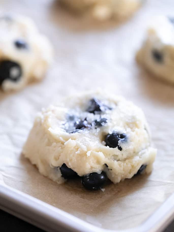 Mounds of white dough with blueberries raw on brown parchment paper lining baking tray