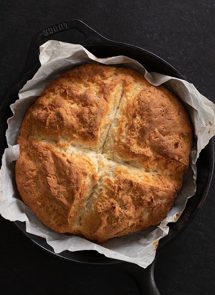 Round brown bread loaf of savory Irish soda bread with cross cut into center in black cast iron pan lined with white paper