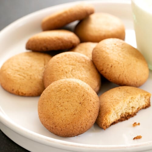 8 gluten free vanilla wafers with one broken on white plate with glass of milk