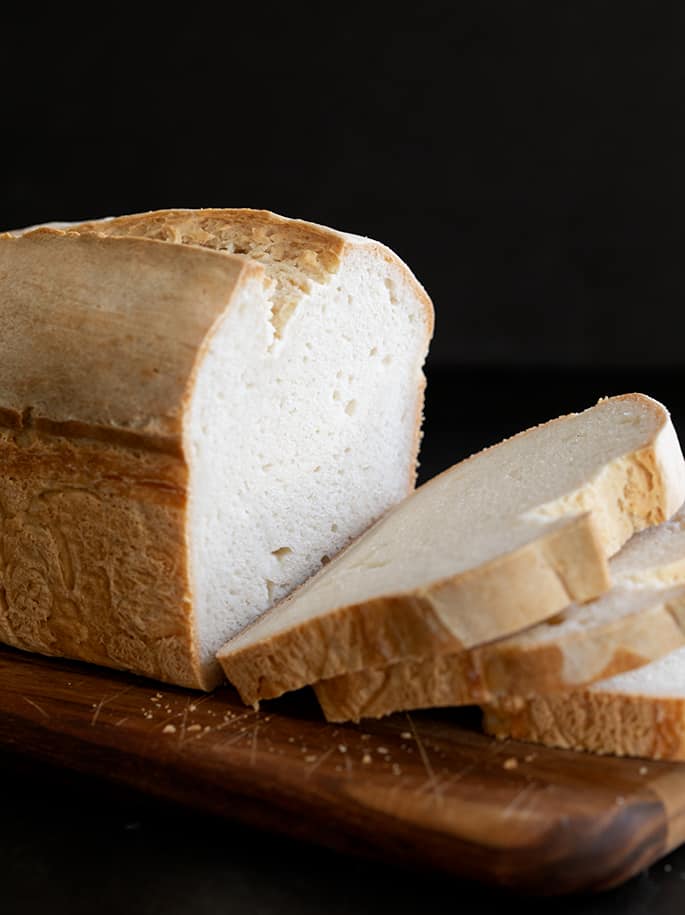 A cut in half white bread bread, sitting on top of a wooden cutting board with crumbs from the cut crust