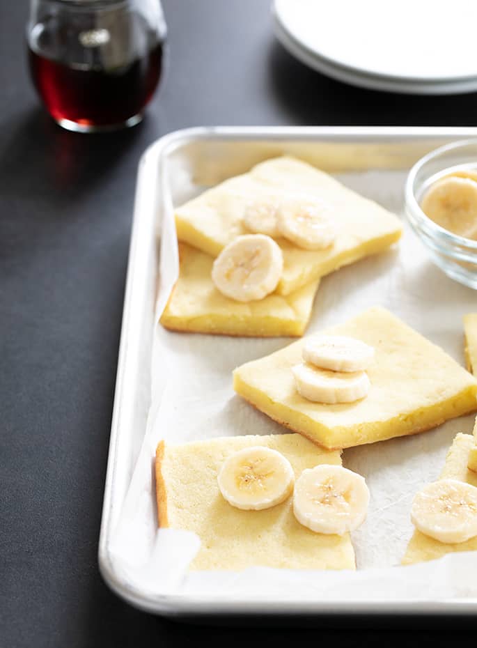 Gluten free pancakes made in a sheet pan, sliced on the baking tray, served with sliced bananas and maple syrup.