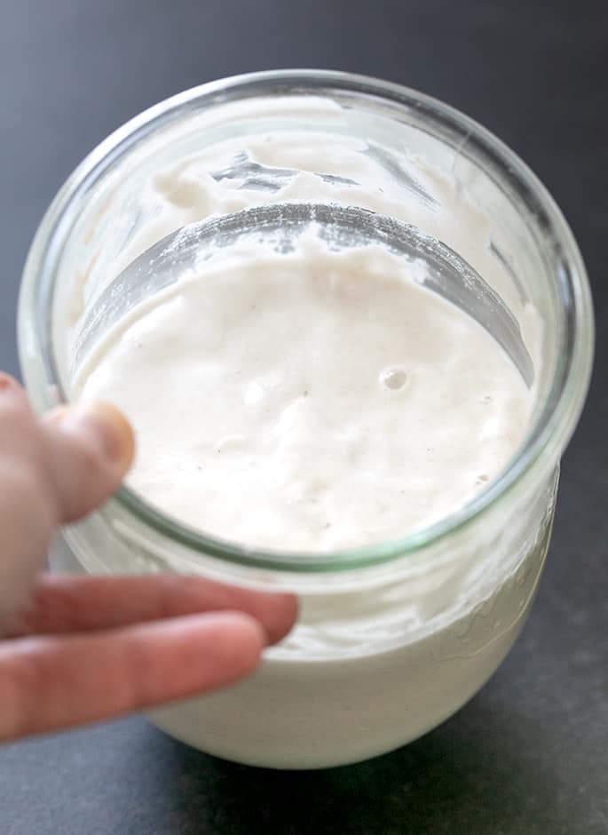 Hand holding glass jar with foamy very light brown liquid showing a rise line after the starter rose overnight.