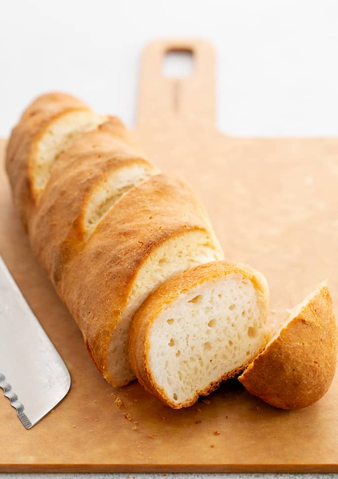 Loaf of gluten free french bread baguette partially sliced on a light brown board with a bread knife