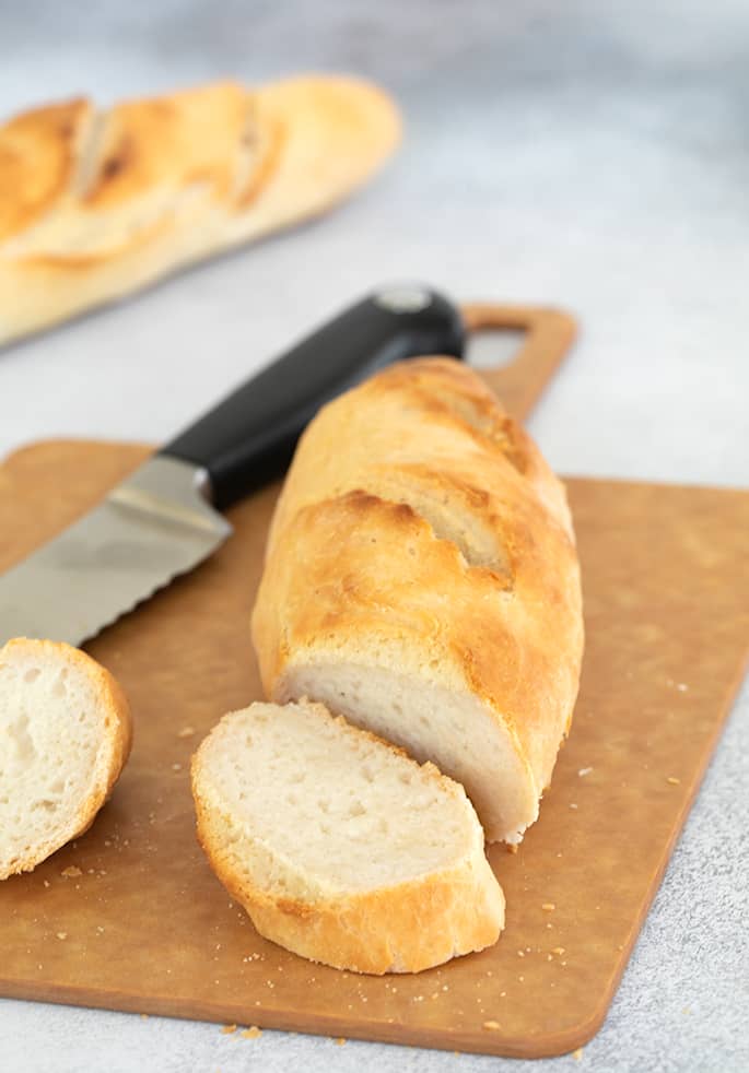 Two loaves of light brown baguette shaped bread, one partially sliced with bread knife on light brown cutting board