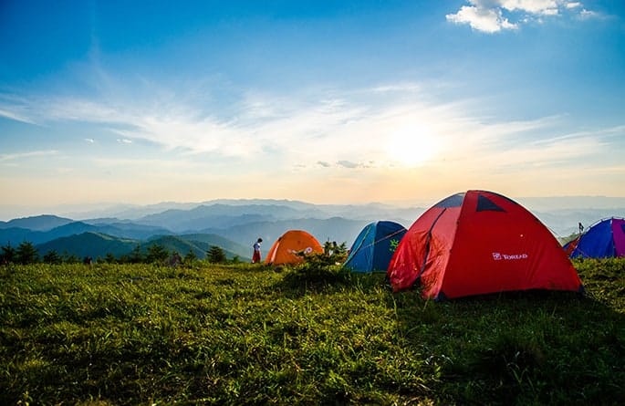 A series of camping tents pitched on a grassy hill overlooking distant misty mountaintops under a sapphire blue sky.