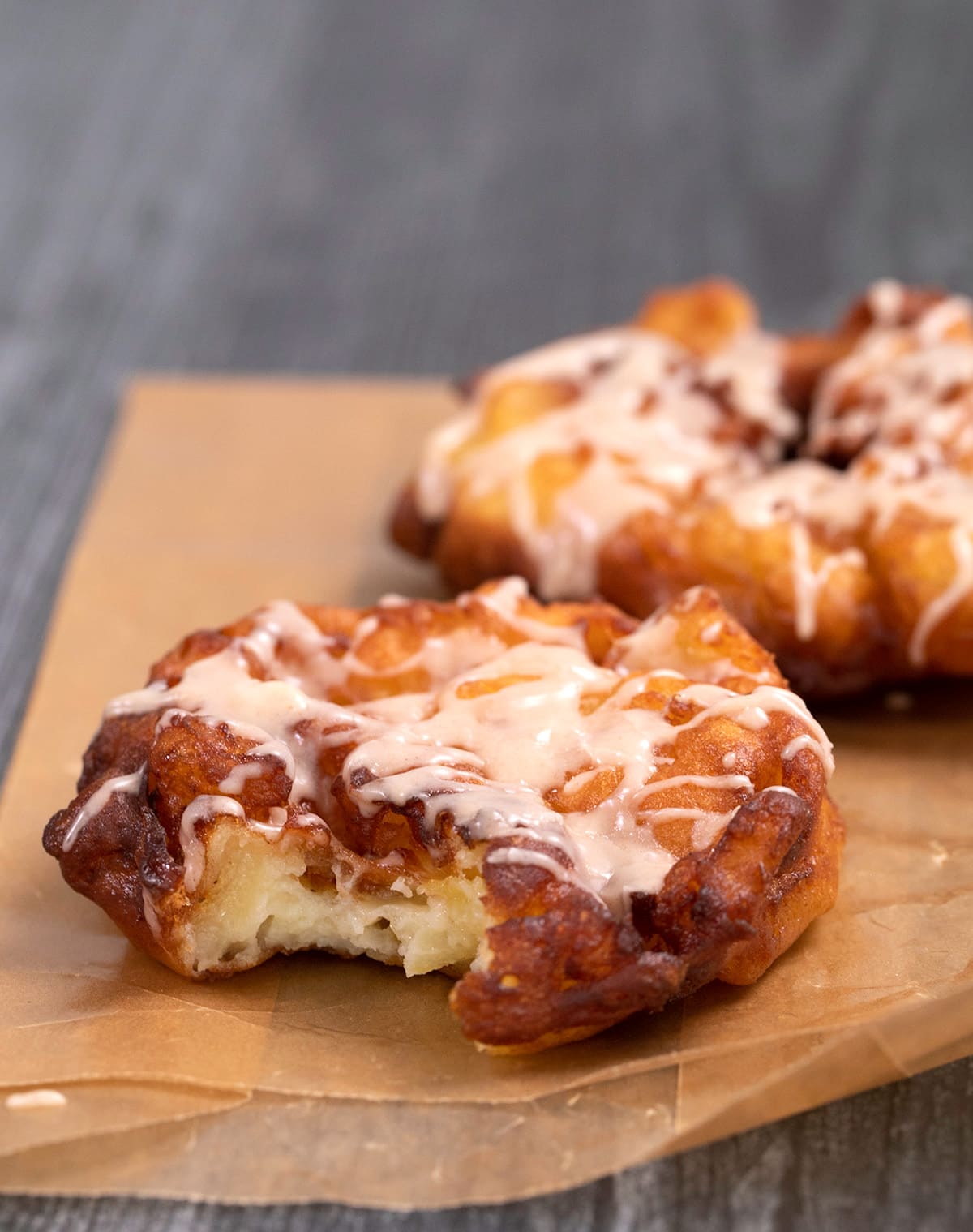 Two gluten free apple fritters with glaze on waxed paper one with a bite taken