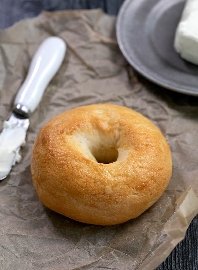 Light brown bagel on brown paper with a knife and cream cheese on a dish