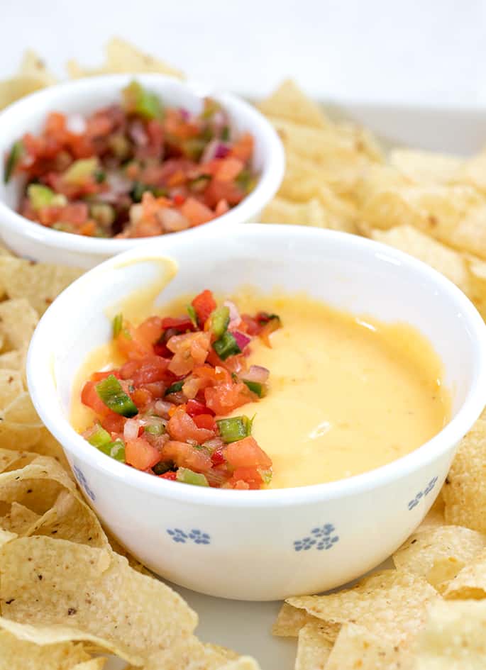 Cheese sauce in white bowl surrounded by chips on white tray