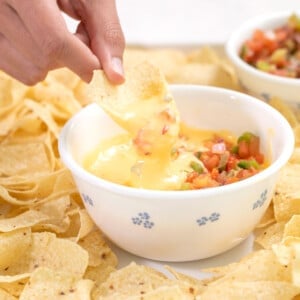 fingers holding tortilla chip and dipping gluten free cheese sauce from small white bowl