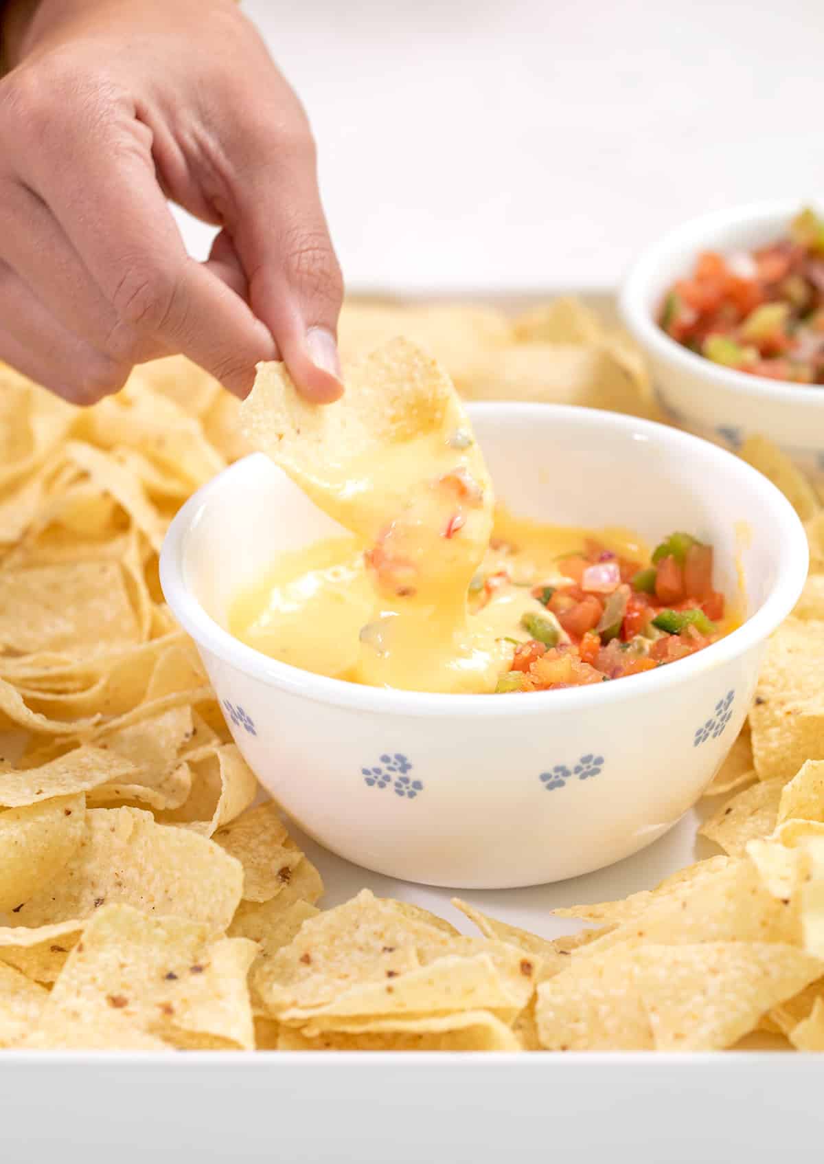 Hand dipping a chip in cheese sauce in a small white bowl with salsa on top