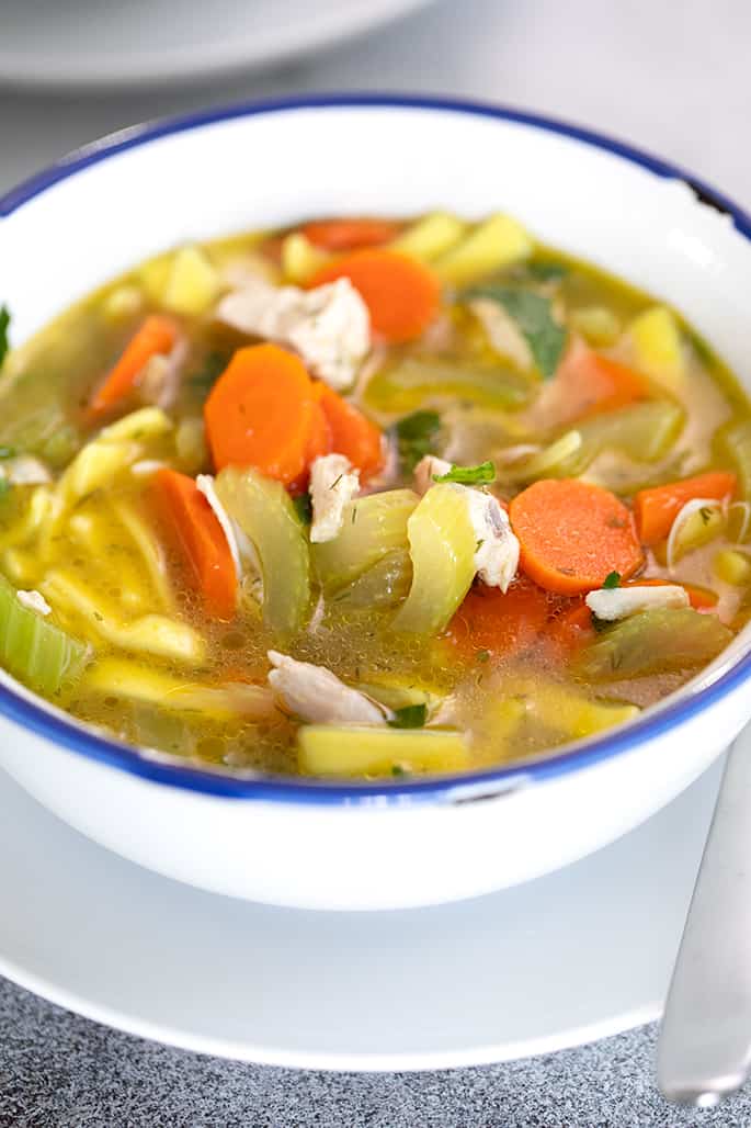 Chicken noodle soup in white bowl with blue rim on white plate with spoon