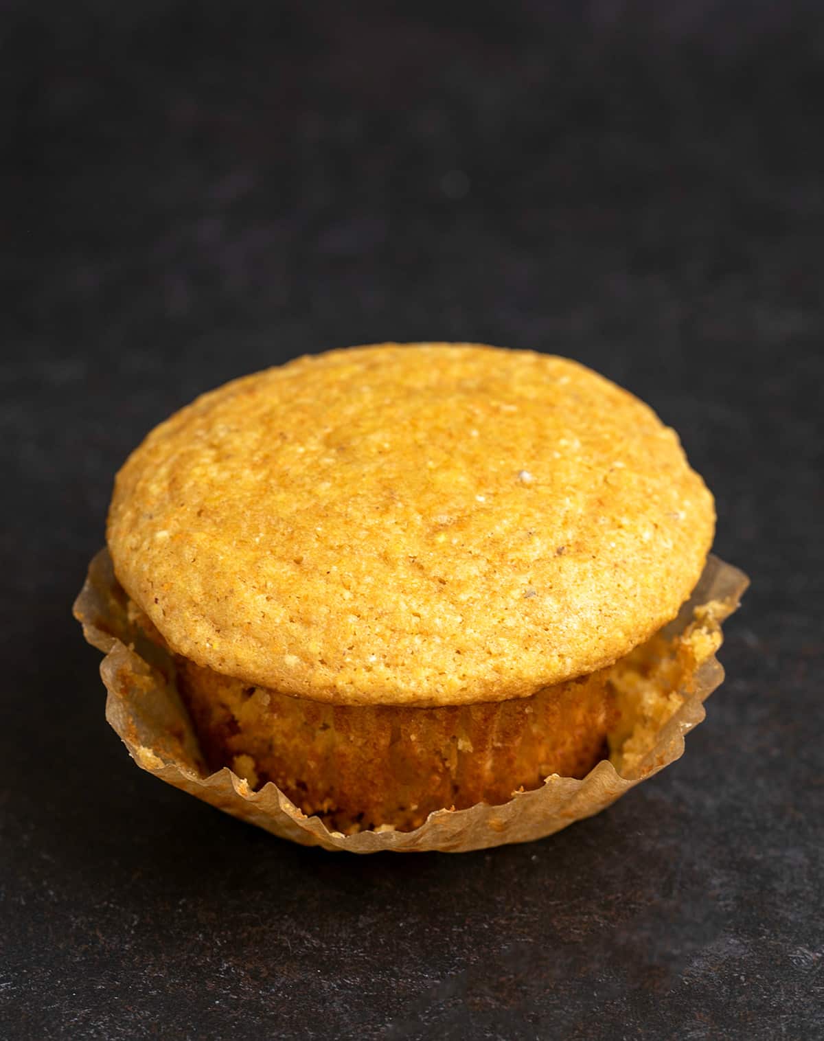 Image of single corn muffin on black surface with brown muffin wrapper pulled down partially