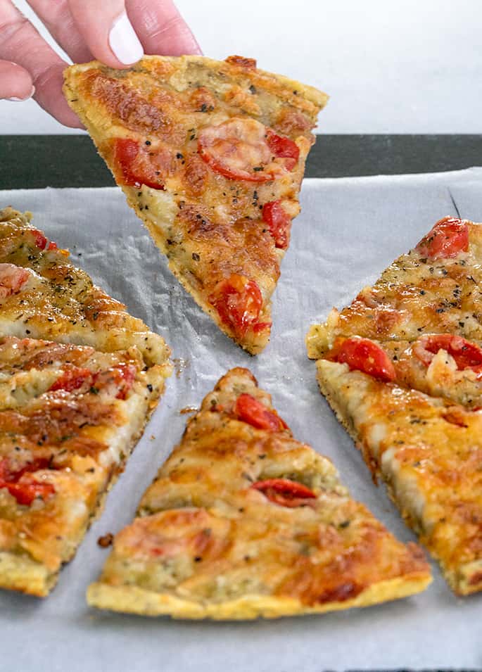 Fingers holding up a slice of chickpea pizza with other slices on white paper