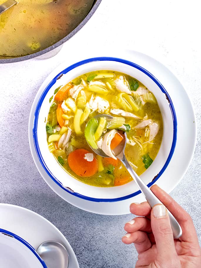 Hand holding spoon with spoonful of chicken noodle soup in white bowl with blue rim