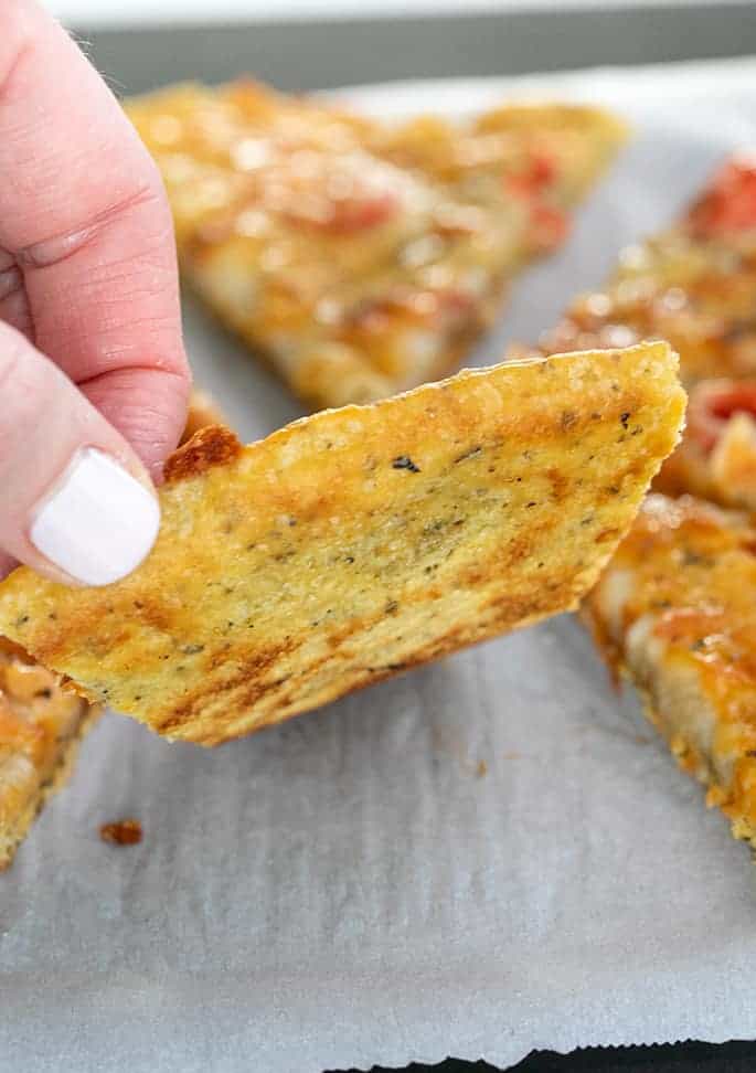 Hand picking up slice of chickpea pizza to show underside of slice on white paper