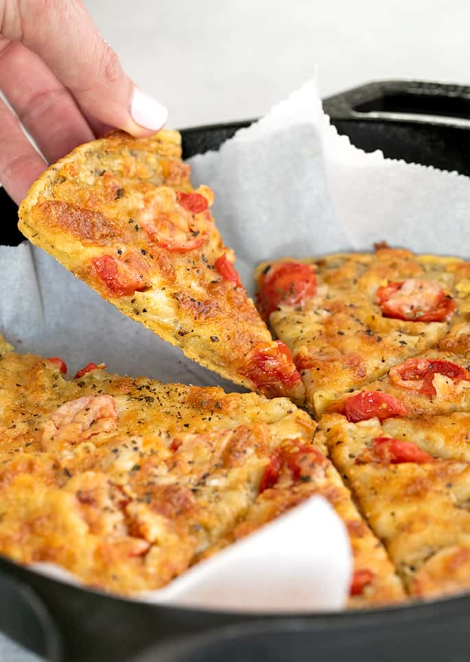 Hand picking up slice of chickpean pizza from cast iron skillet lined with paper