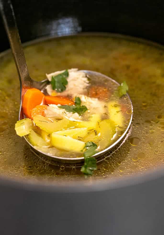 Metal ladle with orange and green vegetables, white chicken pieces, and parsley over a pot of more soup