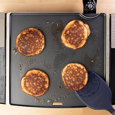 4 cooked banana pancakes on griddle with blue spatula