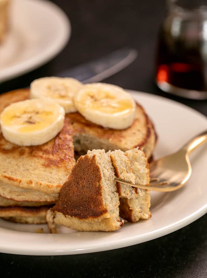 Fork with bite of 3 banana pancakes on a small white plate