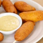 gluten free corn dogs on sticks on white plate with small bowl of yellow mustard