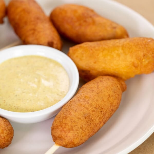 gluten free corn dogs on sticks on white plate with small bowl of yellow mustard