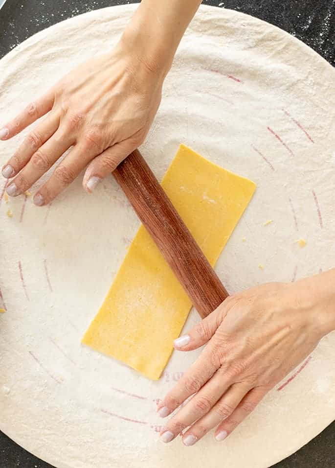 Hands on a small brown rolling pin rolling a piece of pasta dough on a round cloth