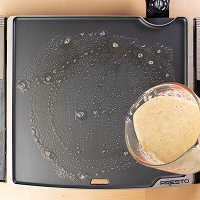 Measuring cup pouring banana pancakes batter onto greased griddle